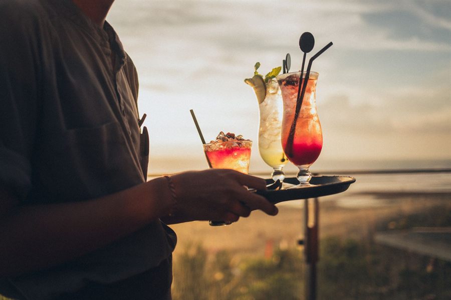Server carrying a tray of colorful cocktails at a seaside bar at sunset