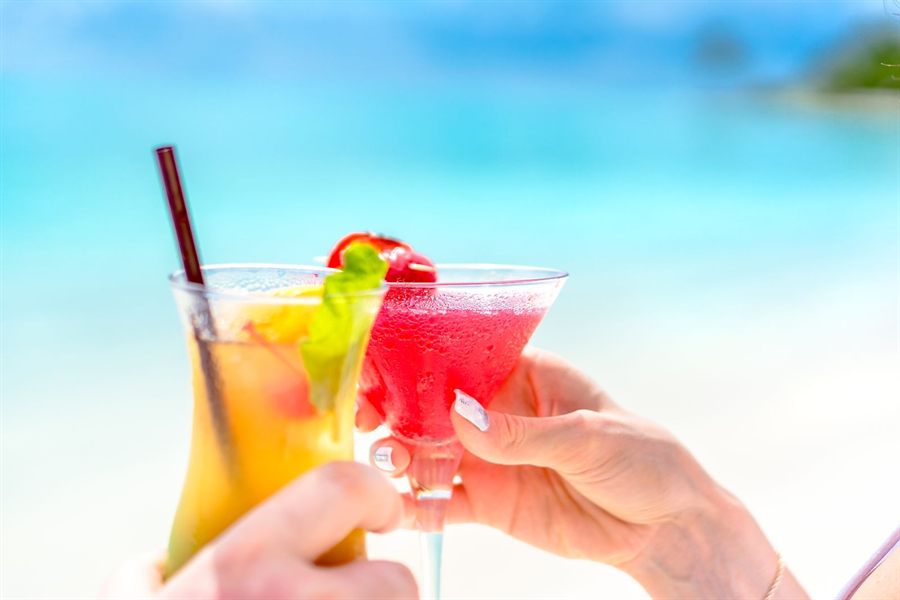 Two hands holding cocktails on a sunny beach with turquoise water